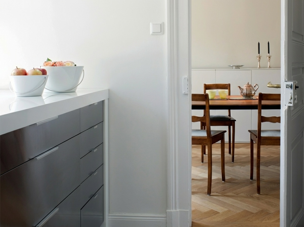 Weißer Küchentresen mit dunkelgrauen Schubladen, darauf zwei weiße Schalen mit roten Äpfeln und rosa Rosen. Eine offene weiße Tür führt zu einem Esszimmer mit Holztisch, drei Holzstühlen, silberner Teekanne und zwei gelb-grünen Gläsern. Der Boden zeigt ein Fischgrätparkett.