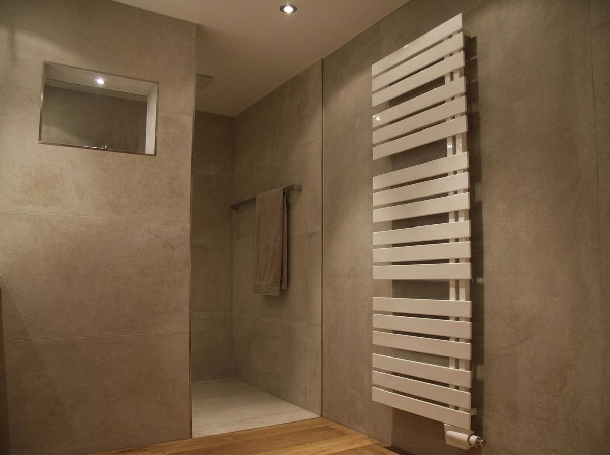 Modernes Badezimmer mit hellbraunen Fliesenwänden, weißem Handtuchheizkörper, begehbarer Dusche mit Glasabtrennung, beigefarbenem Handtuch an der Stange und einem eingelassenen Spiegelschrank über hellem Holzboden.