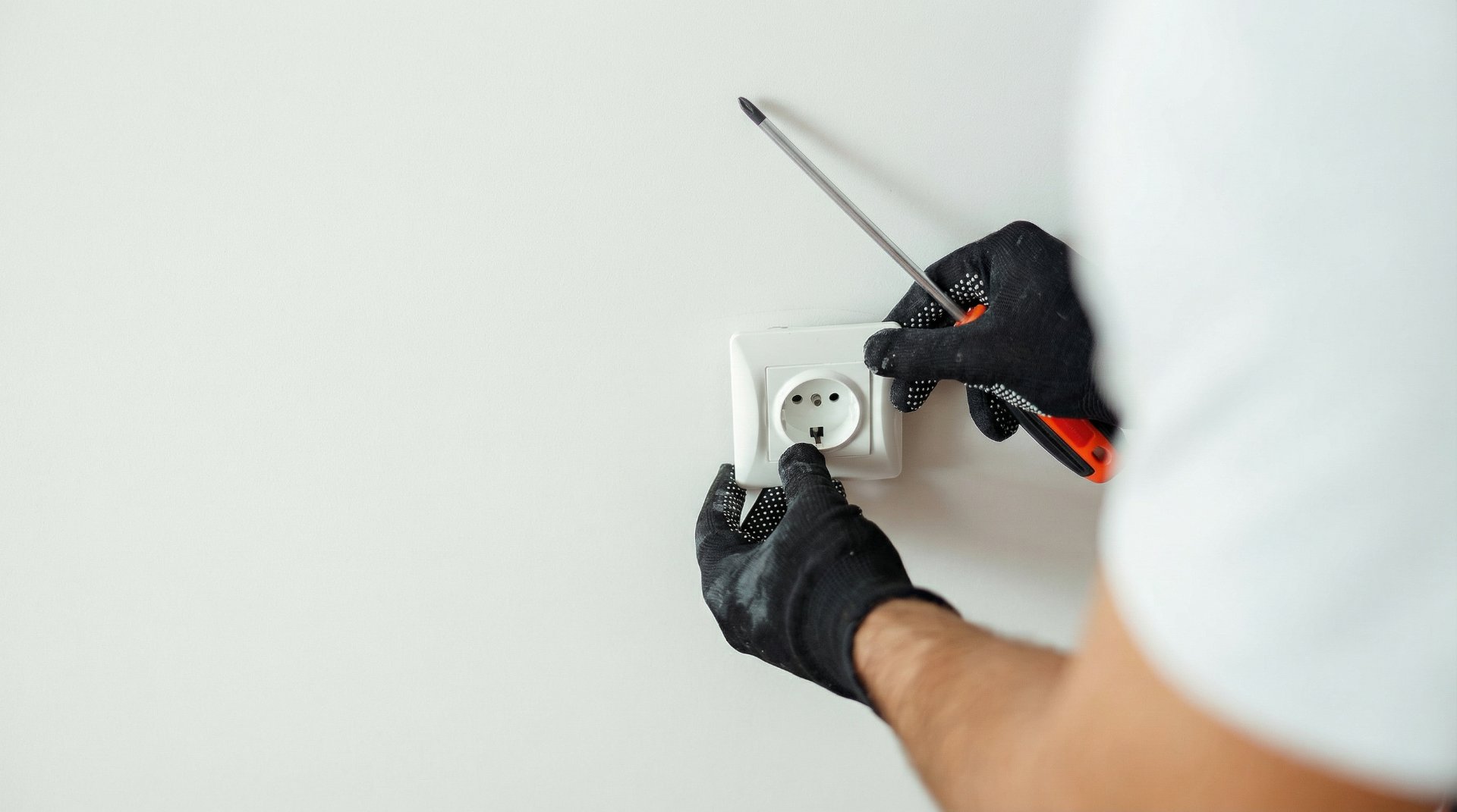 Hände in schwarzen Arbeitshandschuhen montieren eine weiße Steckdose mit einem Schraubendreher an einer weißen Wand, professioneller Service von Graf GmbH.