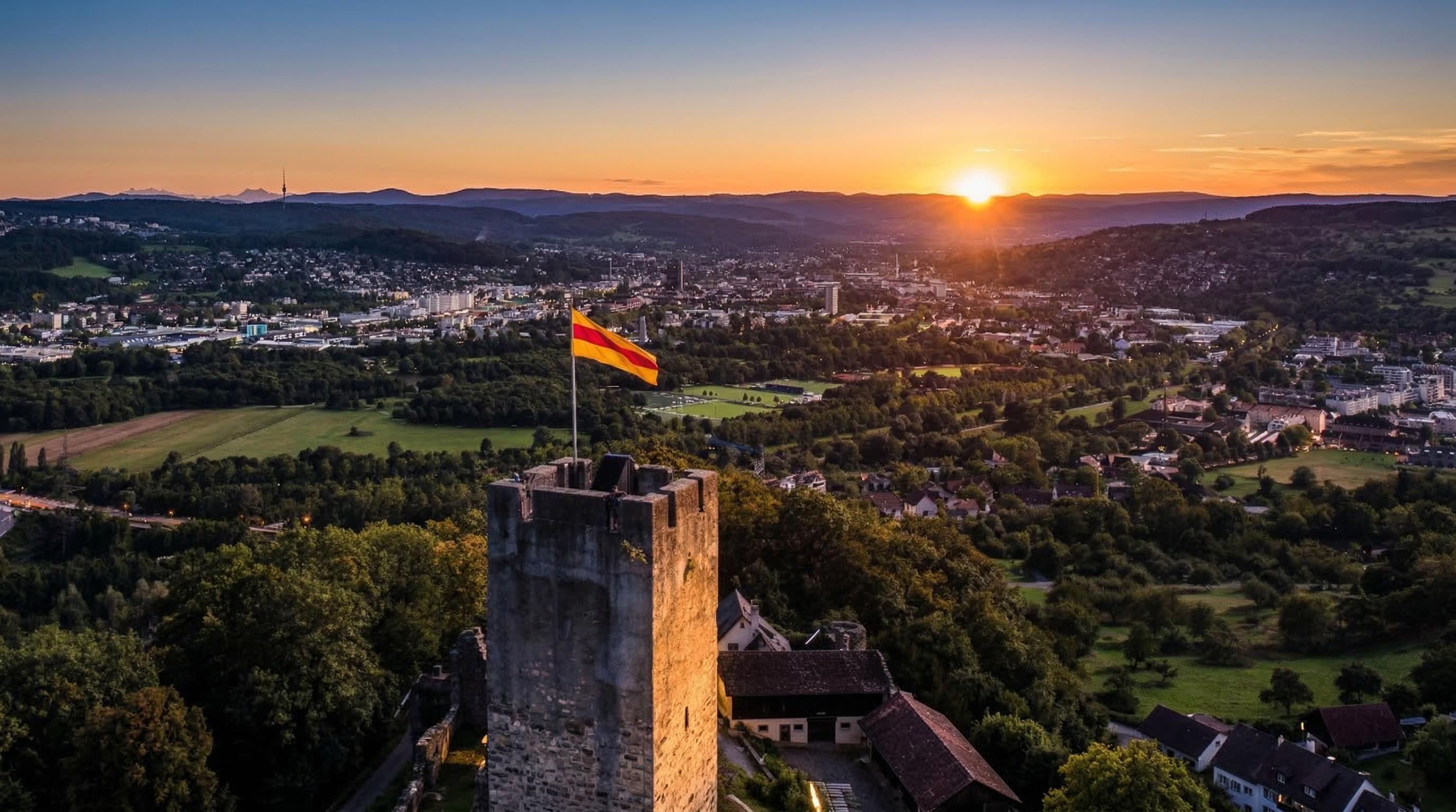 Steinerner Burgturm mit wehender gelb-roter Flagge im goldenen Licht der untergehenden Sonne. Dahinter breitet sich eine Stadt mit Häusern, Wäldern und grünen Feldern in einer hügeligen Landschaft aus. Am Horizont ist die untergehende Sonne sichtbar, die den Himmel orange und blau färbt.