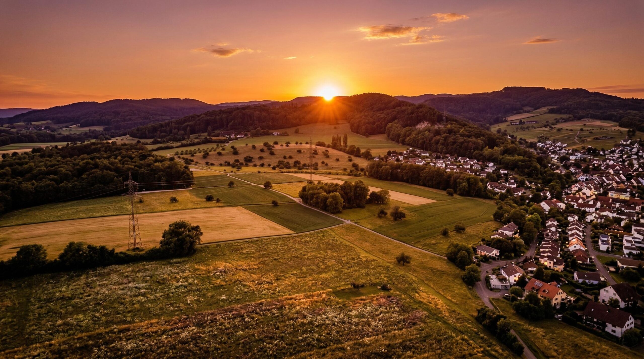 Panoramablick auf eine weite Landschaft bei Sonnenuntergang. Die orangefarbene Sonne strahlt über dunklen, bewaldeten Hügeln am Horizont. Im mittleren und vorderen Bereich erstrecken sich grüne und goldgelbe Felder, dazwischen Baumgruppen und ein Strommast. Rechts ist eine Siedlung mit vielen weißen Häusern und roten Dächern zu sehen, die sich am Hang entlangzieht.