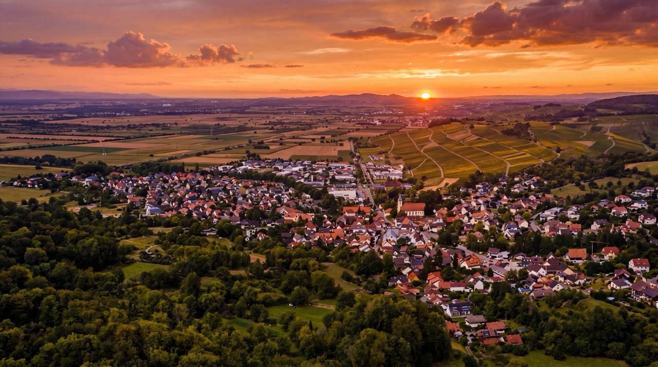 Luftaufnahme von Schliengen mit vielen Häusern und roten Dächern, umgeben von grünen Bäumen, Feldern und terrassierten Weinbergen, bei einem leuchtend orangen und roten Sonnenuntergang über einer hügeligen Landschaft im Hintergrund.