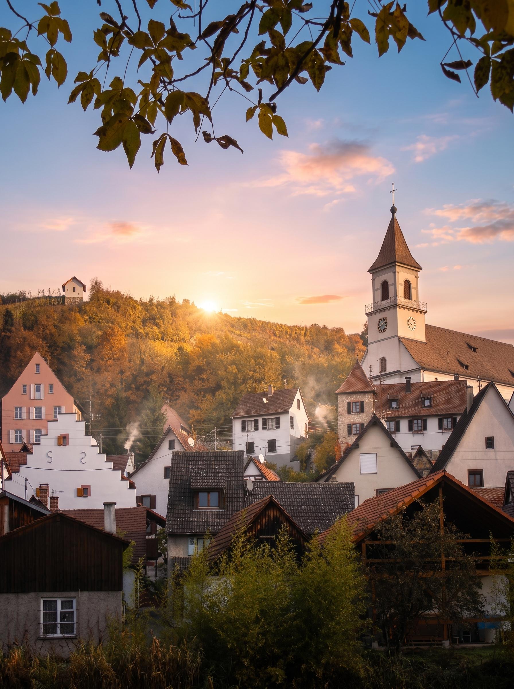 Herbstliche Abendstimmung in einem Dorf in Efringen-Kirchen, geprägt von einer weißen Kirche mit hohem Turm und Glocken. Traditionelle Häuser mit braunen Dächern und aufsteigendem Rauch aus Schornsteinen, die auf funktionierende Heizsysteme von Graf Bad und Heizung Efringen Kirchen hindeuten, säumen den bewaldeten Hang mit goldenen Herbstbäumen. Der Sonnenuntergang taucht den Himmel in warme Orange- und Rosatöne, während grüne Blätter von oben ins Bild ragen.