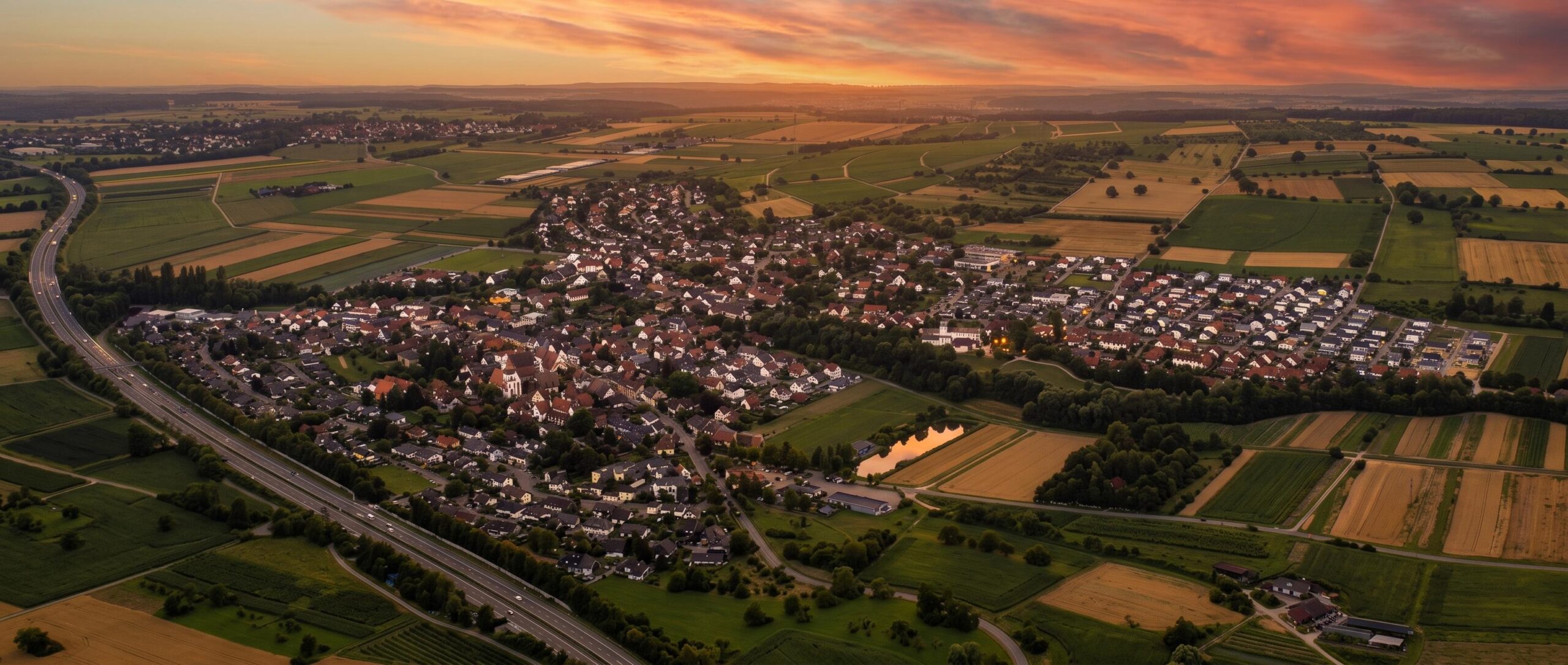 Panoramische Luftaufnahme der Ortschaft Binzen bei Sonnenuntergang, mit zahlreichen Wohnhäusern und roten Dächern, eingebettet zwischen grünen und goldbraunen Feldern. Eine Autobahn mit fahrenden Autos verläuft im linken Bildbereich. Ein kleiner Teich am rechten Bildrand reflektiert die warmen orange-rosa Farben des Abendhimmels, während die ersten Lichter in der Ortschaft Binzen leuchten. Eine idealisierte Darstellung der Umgebung von Graf Bad & Heizung Binzen.
